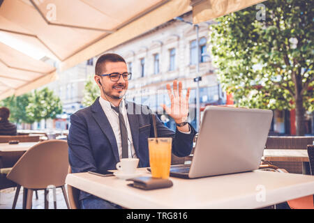 Junge lächelnde Mann an Street Cafe sitzen Winken mit der Hand auf die Kamera unter laptop Beim Videoanruf. Stockfoto