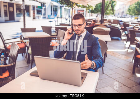 Porträt der jungen Mann auf Handy beim Sitzen an Street Cafe mit einem Laptop. Stockfoto