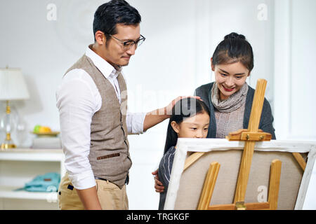 Schöne kleine asiatische Mädchen mit langen schwarzen Haaren und einem Gemälde auf Leinwand, während die Eltern hinter dem Ständigen beobachten. Stockfoto