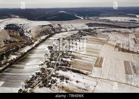 Blick von oben auf die leeren verschneite Felder, Häuser entlang der Straße und bewaldeten Hügeln auf und blauer Himmel. Antenne drone Fotografie, Winterlandschaft. Stockfoto