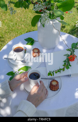 Genießen Kaffee Zeit im Garten. Serviert Tisch mit Blumen, Buch, Kaffee und Dessert im Garten im Frühling. Anordnung mit den Händen einer Frau Stockfoto