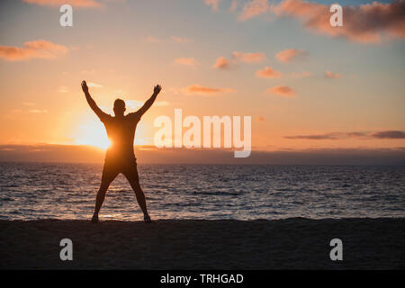 Eine Rückfahrkamera Schuss eine nicht erkennbare Mann tun Jumping Jacks am Strand, er ist mit Blick auf das Meer und den Sonnenuntergang an einem schönen Abend in Stockfoto