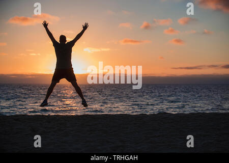 Eine Rückfahrkamera Schuss eine nicht erkennbare Mann tun Jumping Jacks am Strand, er ist mit Blick auf das Meer und den Sonnenuntergang an einem schönen Abend in Stockfoto