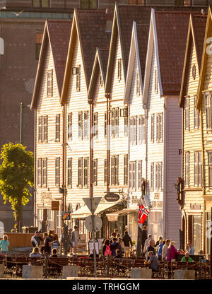 Touristen zu Fuß, andere an den Tischen auf Stühlen vor einem weißen Holzhäuser vorne mit Geschäften im Hafen von Bryggen, Bergen, Hordaland, Norwegen sitzen, Stockfoto