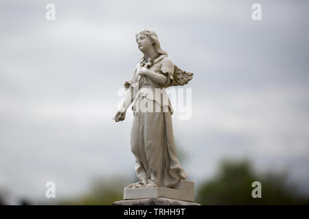 Statue von ein Engel mit einem Kreuz auf der Brust. Der Engel ist ein Teil von einem Grabstein. Im Hintergrund Wolke bedeckte Himmel. Stockfoto