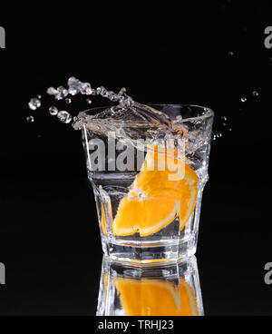 Scheibe der Zitrone Spritzer in ein Glas Wasser mit einem Spray von Wassertröpfchen in der Bewegung in der Luft über dem Glas auf einem dunklen Hintergrund ausgesetzt. Stockfoto