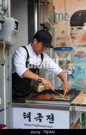 Junger Mann Anbieter kochen Pfannkuchen Buchimgae oder Koreanisch, Koreanisch traditionelles Essen, an einer Garküche in Gamcheon Culture Village in Busan, Südkorea Stockfoto