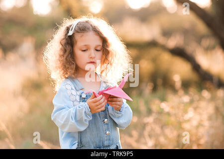 Süße blonde Kind Mädchen 3-4 Jahre alten Holding Papier origami Crane stehen draußen über die Natur Hintergrund. Kinder spielen mit Spielzeug. Kindheit. Stockfoto