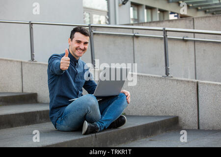 Lächelnd glücklich Mann mit "Daumen hoch" Geste und Laptop auf den Knien, auf einer Treppe Erfolg feiern sitzt. Freelancer Kerl mit Daumen hoch Arbeiten an seinem Hinweis Stockfoto