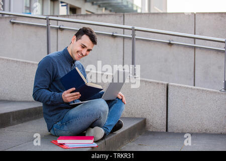 Lächelnd männliche Kursteilnehmer lernen für die Prüfung mit einem Laptop auf den Knien, auf der Treppe sitzen. Lächelnd glücklich Student mit Laptop und lesen Buch tun seiner Pr Stockfoto