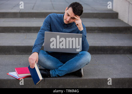 Perplexes Student die Vorbereitung auf eine Prüfung im Campus Bereich sitzen auf der Treppe. Student Mann mit Laptop, sitzen auf der Treppe. Technologie Stockfoto