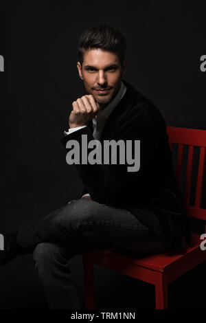 Eine gut aussehende weiße männliche Modell Modellierung in eine formale Kleidung und saß auf einem roten Stuhl Bein gekreuzt, Hand, Kinn, schauen in die Kamera mit Vertrauen. Stockfoto