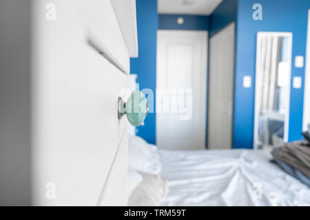Blick in ein Schlafzimmer, einen begehbaren Kleiderschrank, eine Kommode Knopf, blau gestrichenen Wänden, weißen Akzenten und ein Bett ohne Decken, mit unordentlichen, zerknitterten Bettlaken. Stockfoto