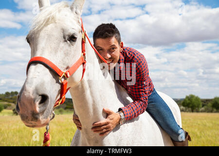 Neugierige junge Kerl in casual Outfit lächelnd und umarmt den Hals des weißen Pferd während der Fahrt in das Feld An bewölkten Tag Stockfoto