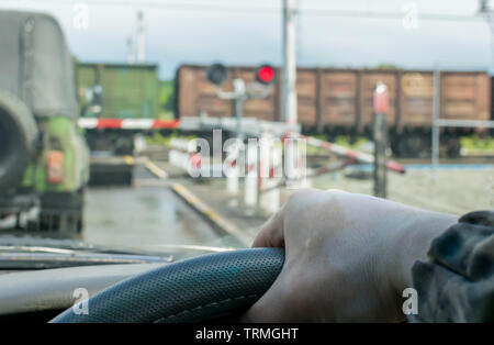 Blick auf die Fahrer Hand am Lenkrad des Autos, die vor einem geschlossenen Bahnübergang an einer roten Ampel angehalten. Der Fahrer wartet Stockfoto