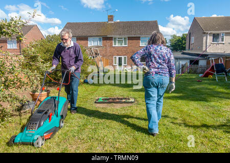 Ein rentnerehepaar, Gartenarbeit in einem Wohngebiet zurück Garten an einem sonnigen Tag mit blauen Himmel. Der Mann mäht den Rasen mit einem elektrischen Rasenmäher. Stockfoto