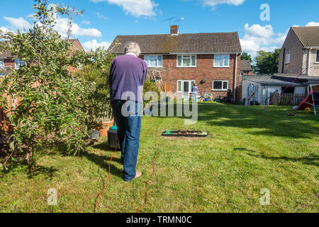 Ein Mann mäht den Rasen in einem Wohngebiet zurück Garten mit einem elektrischen Rasenmäher an einem sonnigen Tag mit blauen Himmel im Frühsommer. Stockfoto