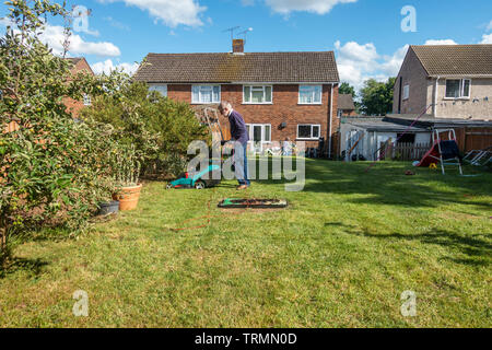 Ein Mann mäht den Rasen in einem Wohngebiet zurück Garten mit einem elektrischen Rasenmäher an einem sonnigen Tag mit blauen Himmel im Frühsommer. Stockfoto