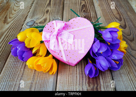 Rosa Geschenkverpackung in Form eines Herzens, mit weißen Bändern mit Bogen gebunden, und Blumenstrauß aus gelb und lila Krokusse Blumen sind auf eine unlackierte Holz- s Stockfoto