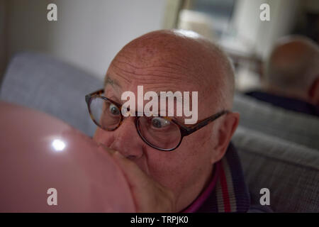 Ältere Menschen im Ruhestand mann Blasen einer großen herzförmigen Rosa Ballon das Zimmer für eine Geburtstagsfeier zu verzieren Stockfoto