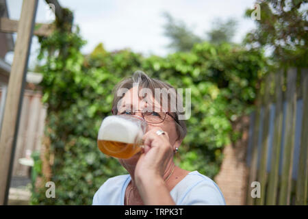 Glückliche und Gesunde ältere Frau trinkt ein großes Glas Bier draußen im hellen Sommer Sonnenschein Stockfoto