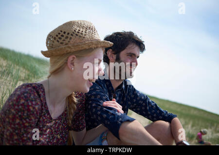 Schöne junge Paare sitzen auf einem Hügel auf einer Düne genießen die Aussicht und die frische Meeresbrise am Strand in hellen Sommer Sonnenschein Stockfoto