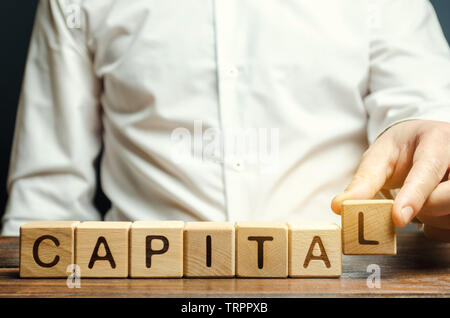 Ein Mann legt Holzblöcke mit dem Wort Hauptstadt. Die Kombination von materiellen, intellektuellen und finanziellen Ressourcen, die für die Gewinne verwendet, zusätzliches Material Stockfoto