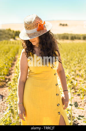 Porträt der jungen Frau mit dem Lockigen schwarzen Haar, gelben Kleid und Stroh Hut mit Blume im natürlichen Platz bei Sonnenuntergang. Frau Blick in die Landschaft. Stockfoto