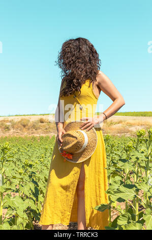 Nicht erkennbare Frau mit gekräuselte schwarze lange Haare, gelben Kleid und Stroh Hut in der farbenfrohen Landschaft. Frau im ländlichen Bereich an einem sonnigen Tag. Stockfoto