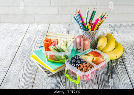 Gesunde Schule Lunch Box. Kid's Lunch Box mit Sandwiches, Obst, Gemüse, Nüssen, Wasser- und Schulbedarf, weiß Holz- Hintergrund Stockfoto