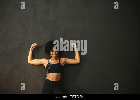 Starke Frau flexing Muskeln und lächelnd gegen graue Wand. Afrikanische weibliche model in Sportkleidung ihre Muskeln n Gym angezeigt. Stockfoto