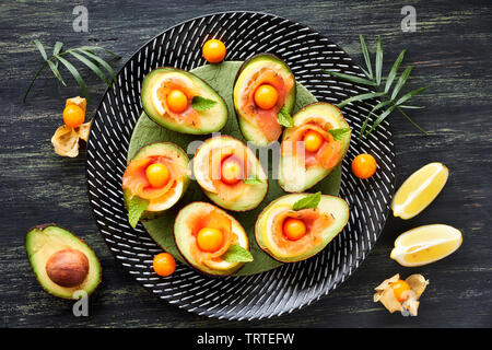 Avocado Boote mit geräuchertem Lachs, Zitrone, Physalis und Minzeblatt, Ansicht von oben auf dunkel strukturierten Hintergrund Stockfoto