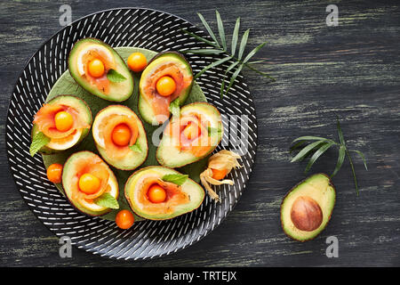 Avocado Boote mit geräuchertem Lachs, Zitrone, Physalis und Minzeblatt, Ansicht von oben auf dunkel strukturierten Hintergrund Stockfoto