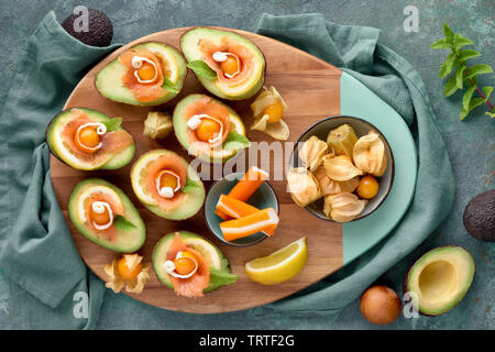 Avocado Boote mit geräuchertem Lachs, Zitrone, Physalis und Minzeblatt, Ansicht von oben auf Blau-grün strukturierten Hintergrund Stockfoto