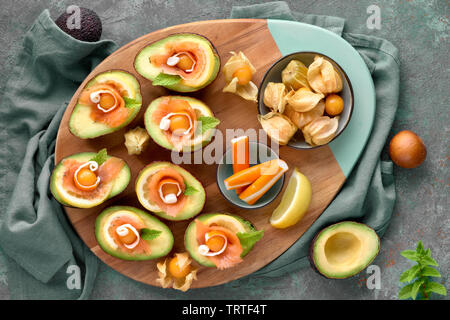 Avocado Boote mit geräuchertem Lachs, Zitrone, Physalis und Minzeblatt, Ansicht von oben auf Blau-grün strukturierten Hintergrund Stockfoto