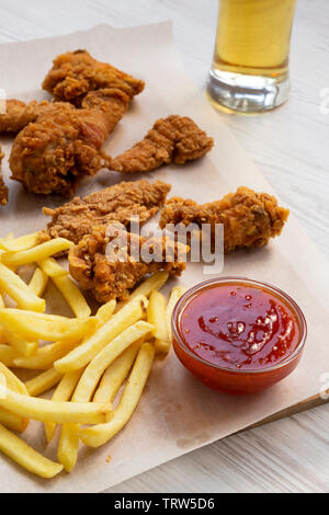 Lecker Fastfood: Fried Chicken Drumsticks, würzige Flügel, Pommes frites und Chicken Strips mit Sauer-süßen Soße und kaltes Bier auf einem weißen Holztisch, lo Stockfoto