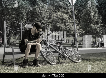 Ein junger Mann mit einem tätowierten Arm, Kopfhörer in den Ohren, sitzt auf einer Bank im Park und zieht sein Handy. Neben ihm und seinem Fahrrad. Entsättigung. Stockfoto