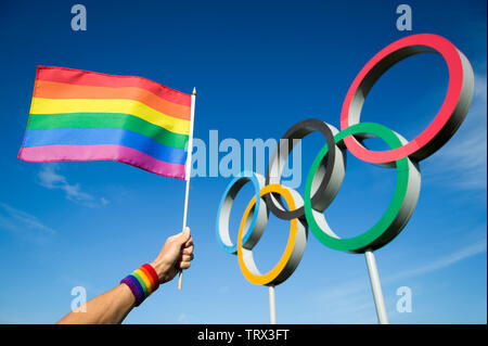 LONDON - Mai 4, 2019: eine Hand regenbogenfarbenen Schweißband Wellen tragen ein Gay Pride Flagge hängt vor der olympischen Ringe gegen den strahlend blauen Himmel. Stockfoto