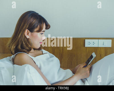 Schöne asiatische Frau lächelnd auf dem Bett und Sie ihr Smartphone auf der weißen Wand und Holz- Hintergrund mit Kopie Platz im Hotel Schlafzimmer. Stockfoto