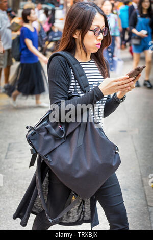 Asiatische Frau konzentriert sich auf mobiles Gerät, während der Überquerung der Straße in Hong Kong, SAR, China Stockfoto