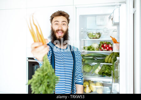 Porträt von einem gut aussehenden Mann mit Karotte in der Nähe der Kühlschrank voll mit frischem Obst und Gemüse zu Hause stehend Stockfoto