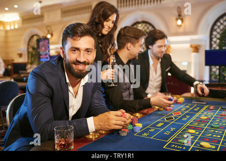Eine junge Brünette bärtiger Mann im Anzug mit einem Glas Whiskey in der Hand sitzen am Tisch Roulette Poker Spielen in einem Kasino. Stockfoto