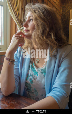 Eine schöne, gut gekleidete junge Frau, 23 Jahre alt, mit Lange wellige Haare, allein sitzen und schaut nachdenklich aus dem Fenster. Stockfoto