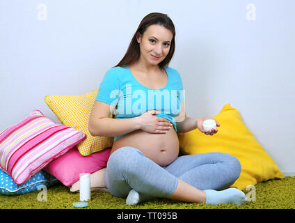 Junge regnant Frau sitzt auf dem Boden und die Sahne auf ihrem Bauch auf Wand Hintergrund Stockfoto