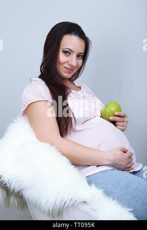 Junge regnant Frau sitzt auf einem Sessel und mit Apfel auf Wand Hintergrund Stockfoto