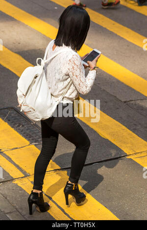 Chinesisches Mädchen konzentriert sich auf Handy Überqueren der Straße, Hong Kong, SAR, China Stockfoto