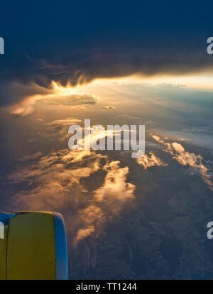 Dramatische Sonnenstrahlen brechen durch Sturmwolken, die von einem Flugzeugfenster aus gesehen werden, mit Flügel und Motor über der glühenden Wolkenlandschaft. Stockfoto