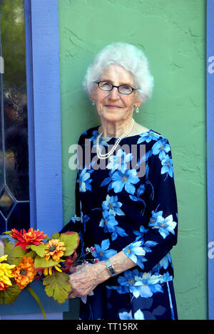 Klassische ältere Frau arrangiert Blumen in einem Fenster außerhalb ihrer Geschenke Shop. Sie trägt ein Kleid und Perlen und einem leichten Lächeln. Stockfoto
