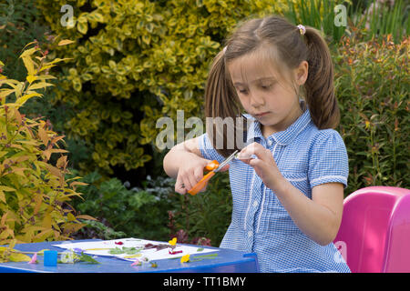Acht Jahre alten Mädchen in Schuluniform Kleid in den Garten, macht ein Bild von Blumen und Blättern auf Papier geklebt, Natur, Kunst, Handwerk, mit einer Schere Stockfoto