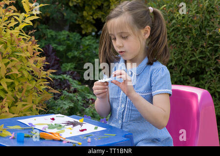 Acht Jahre alten Mädchen in Schuluniform Kleid in den Garten, macht ein Bild von Blumen und Blättern auf Papier geklebt, Natur Kunst Handwerk Stockfoto